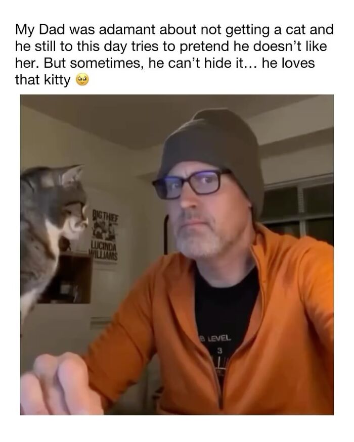 Man in an orange jacket and beanie looking at a cat unaware of how goofy the cat is in a cozy indoor setting.