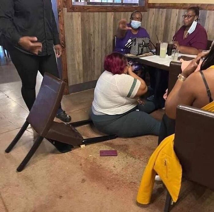 Woman sitting on the floor with a broken chair under her at a restaurant, others seated at a table nearby, exorcist theme.