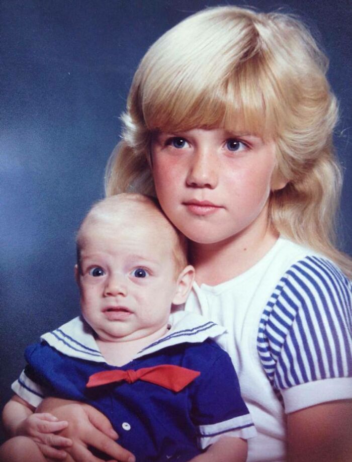 Niña rubia con camiseta a rayas sostiene a bebé con expresión incómoda en fotografía familiar icónica y curiosa.