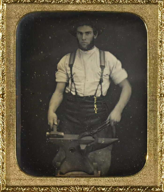 Blacksmith posing with tools and an anvil in a vintage photo from the time when early photography was a new phenomenon.