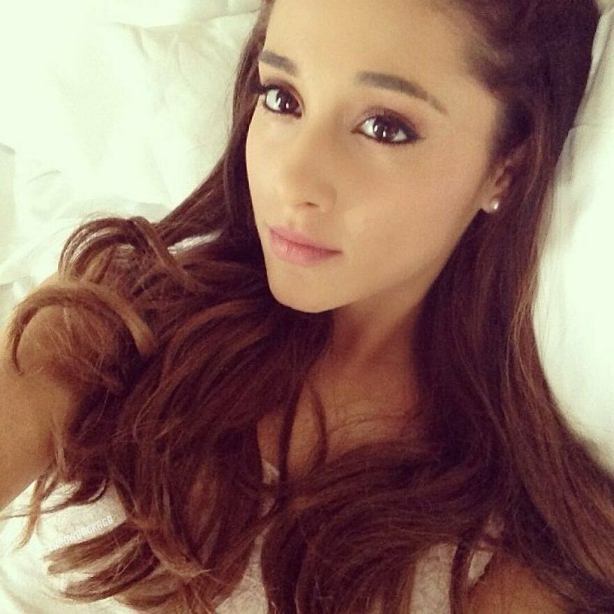 Ariana Grande close-up selfie showing her smooth skin and long brown hair in a relaxed pose on a white pillow.