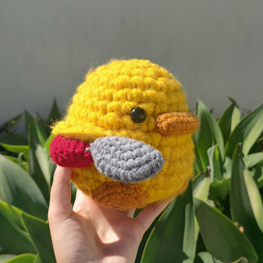 Hand holding a bright yellow crochet animal resembling a duck with gray and red wings and an orange beak outdoors.