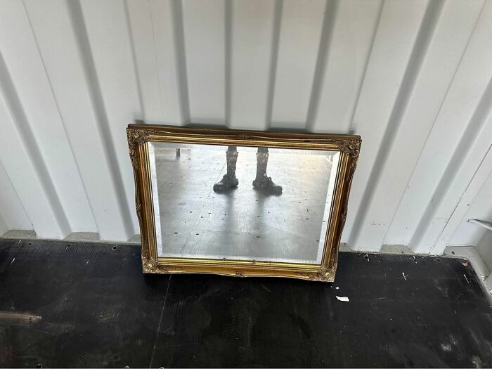 Vintage mirror with ornate gold frame reflecting a person's boots on a black floor against a white wall.