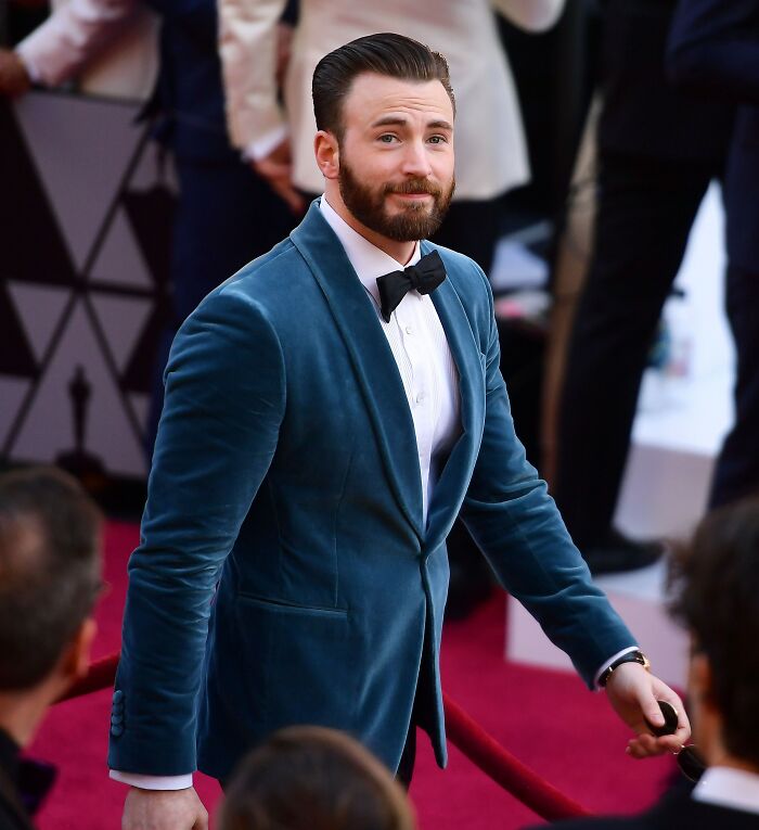 Chris Evans in a blue velvet tuxedo jacket and bow tie attending a red carpet event with fans around him.