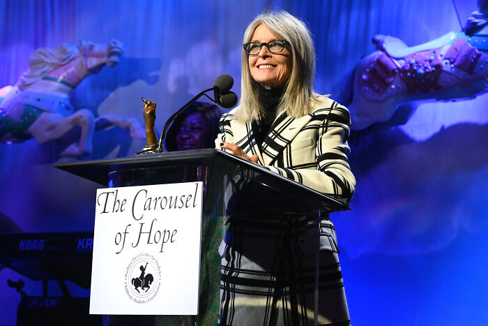 Diane Keaton speaking at a podium with The Carousel of Hope banner, smiling and wearing glasses and a striped jacket.