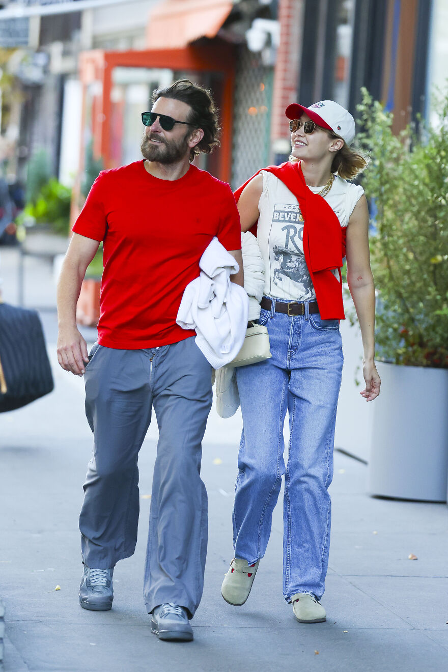 Gigi Hadid and Bradley Cooper walking casually on city street, both wearing sunglasses and smiling together.
