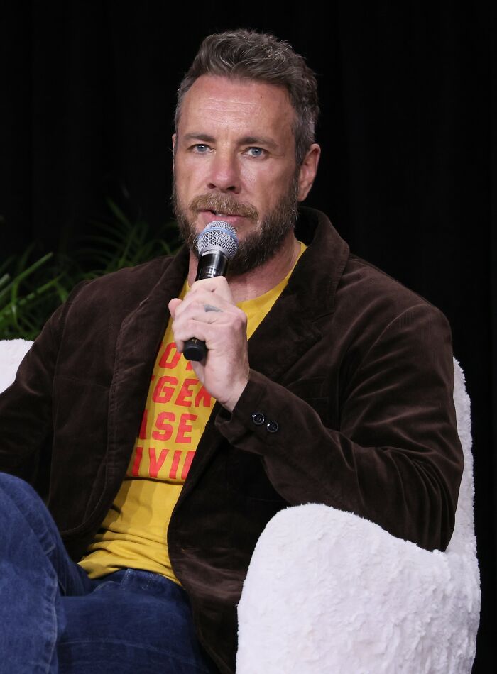 Man with beard speaking into a microphone, seated in a white chair, related to Kristen Bell snubs fans controversy.