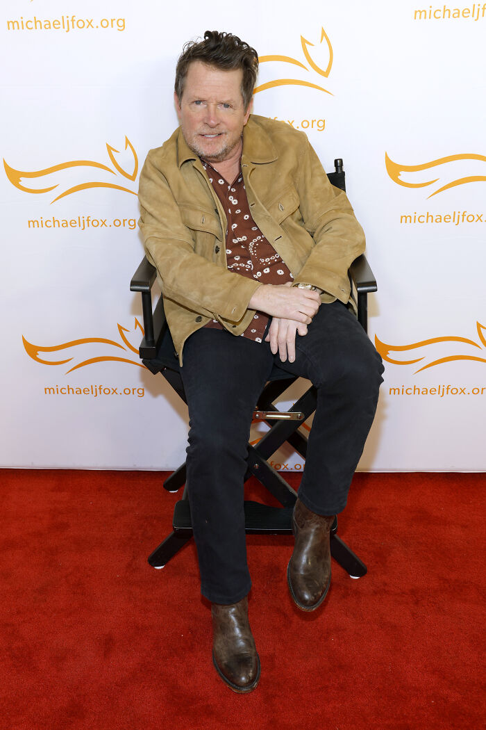 Michael J. Fox sitting in a chair at a michaeljfox.org event, wearing a tan jacket and brown boots, discussing health update.