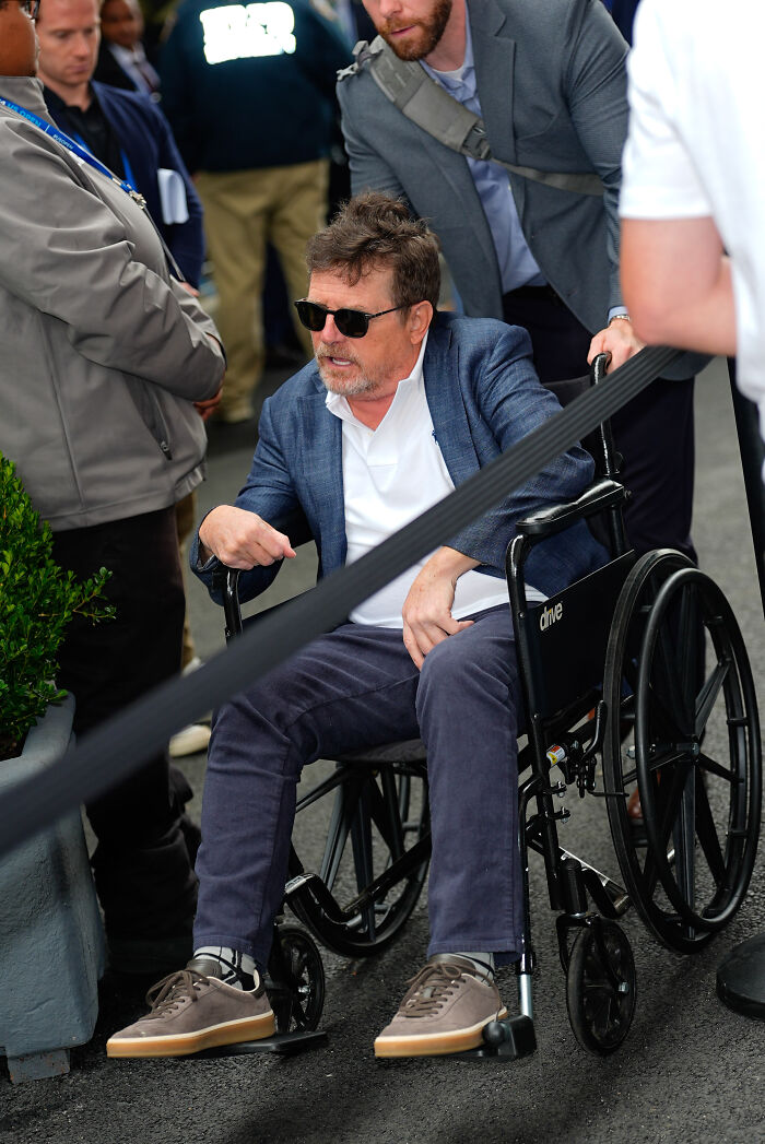 Michael J. Fox in a wheelchair wearing sunglasses and a blazer, sharing a heartbreaking health update with a crowd nearby.
