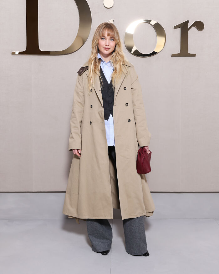 Jennifer Lawrence at Paris Fashion Week wearing a beige trench coat and gray pants in front of Dior logo.