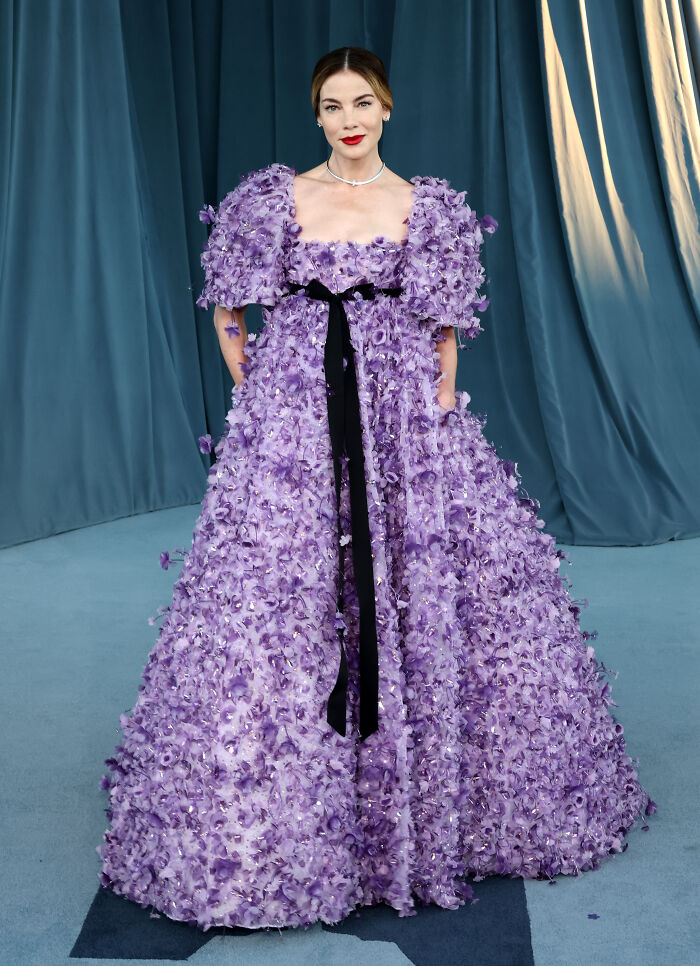 Woman in a detailed lavender gown with floral appliques and puff sleeves on the Academy Museum Gala red carpet.
