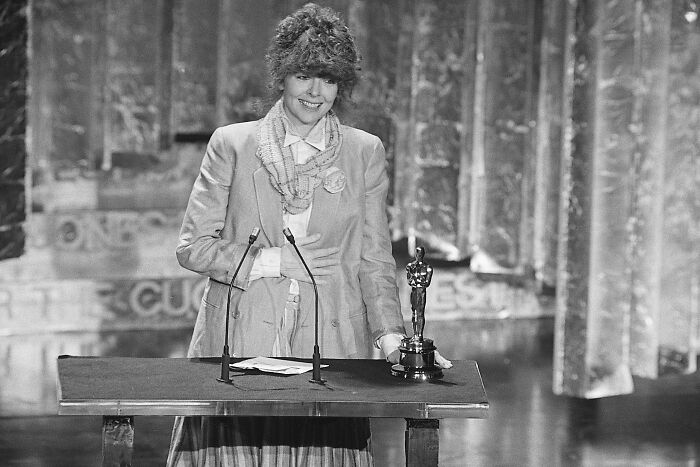 Diane Keaton smiling at a podium while holding an award, spotlight on her during a formal event.