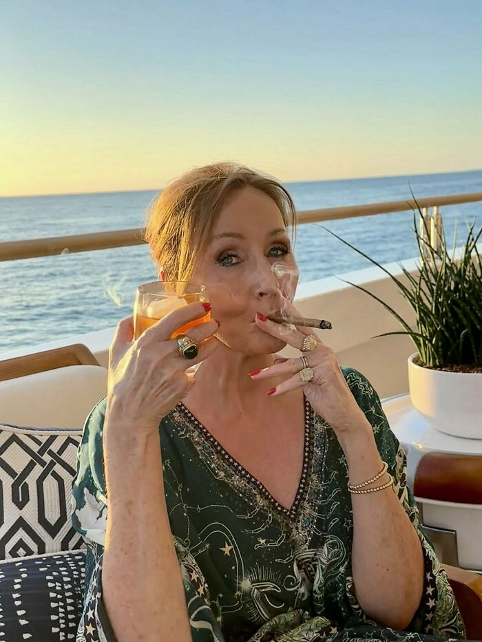 Woman smoking a cigar and holding a drink by the ocean, related to J.K. Rowling and trans rights discussion.