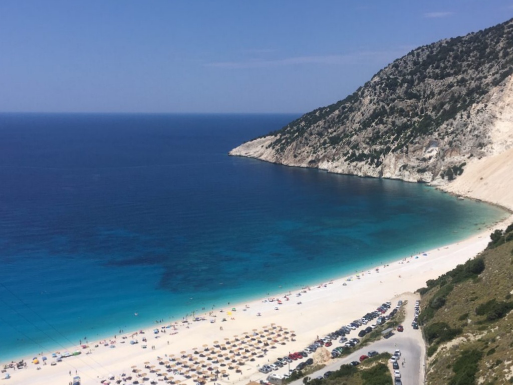 Aerial view of a coastal beach with clear blue water, umbrellas, and parked cars along the shore.