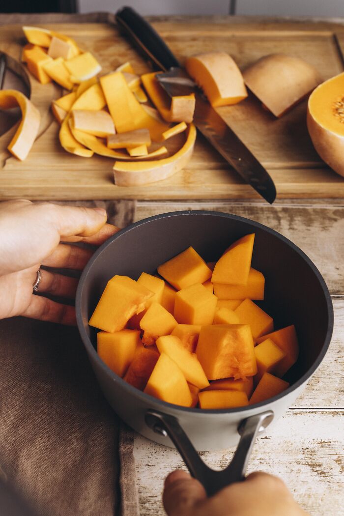 Chopped butternut squash in a pot with peels and knife on wooden cutting board for autumn cake recipe.