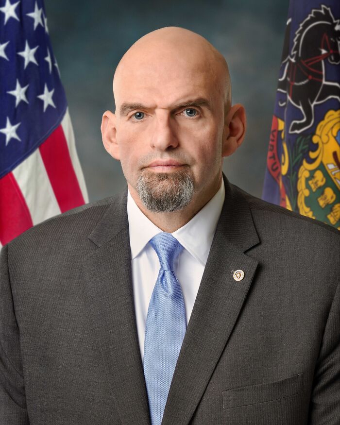 Bald man in a suit with a blue tie posing formally in front of American and state flags, symbolizing disappointment in someone idolized.