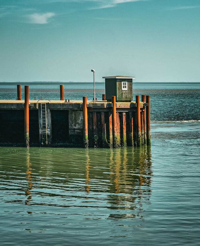 Minimalist architectural vision shown in a simple pier structure with a small building by calm waters and clear sky.