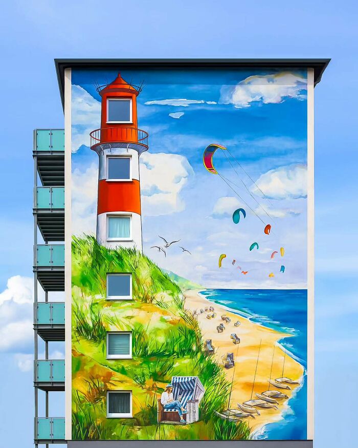 Mural on minimalist architectural building featuring lighthouse, beach scene, and colorful kites against vibrant blue sky.