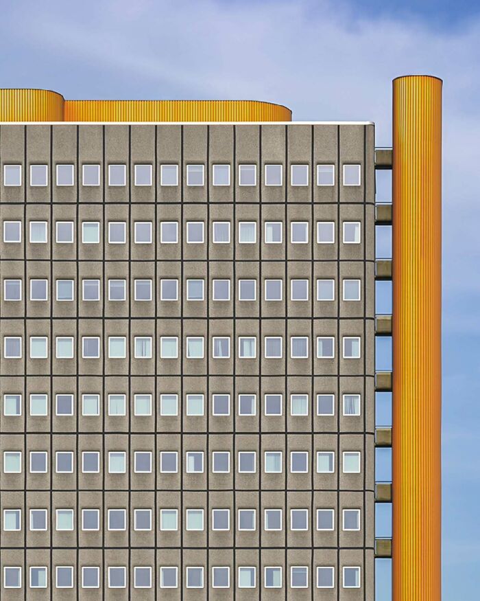 Minimalist architectural vision of Olle L shown in a grid-patterned building facade with yellow cylindrical accents against a blue sky.