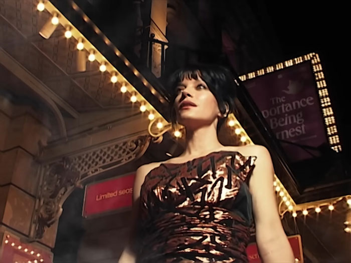 Lily Allen standing outside a theater at night, spotlighted with lights, related to brutal album about David Harbour.