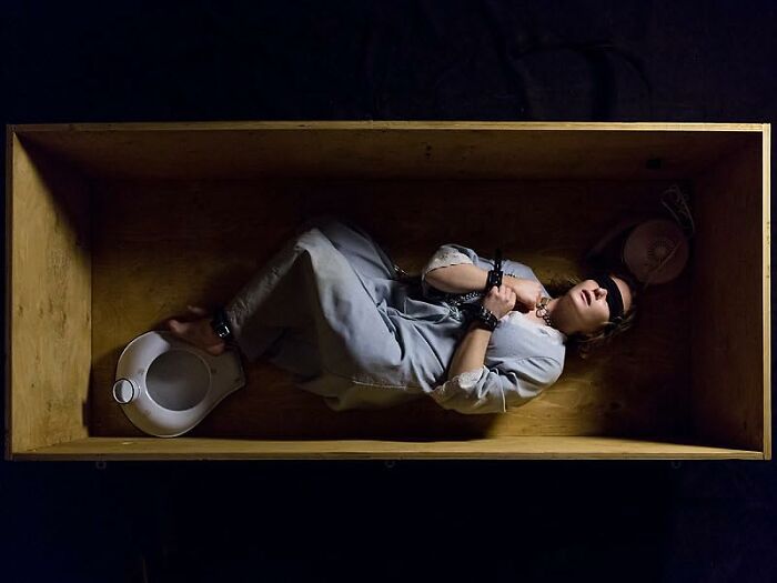 Person in gray clothing lying blindfolded and handcuffed inside a wooden box, evoking movies based on true stories.