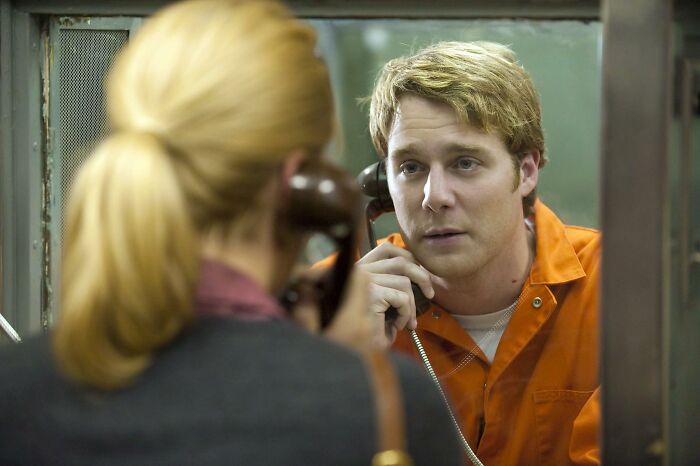Man in an orange prison jumpsuit talking on a prison phone through a glass barrier in a movie based on true stories