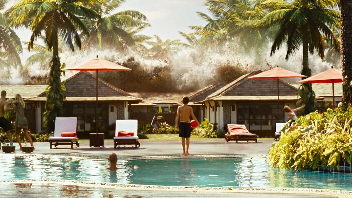 Large ocean wave crashing behind tropical resort pool with people reacting to the scene in a movies based on true stories.