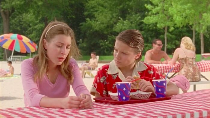 Two young people sitting at a picnic table outdoors with drinks, scene from movies based on true stories.