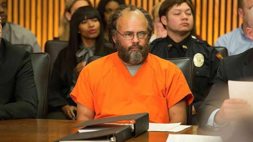 Middle-aged man in an orange prison jumpsuit sitting in a courtroom, a scene from movies based on true stories.