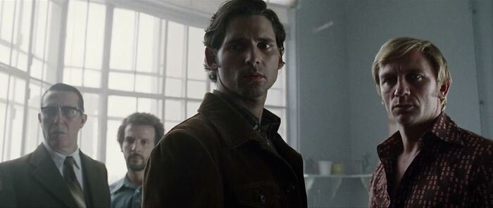 Four men in 1970s attire standing in a dimly lit room, evoking scenes from movies based on true stories.