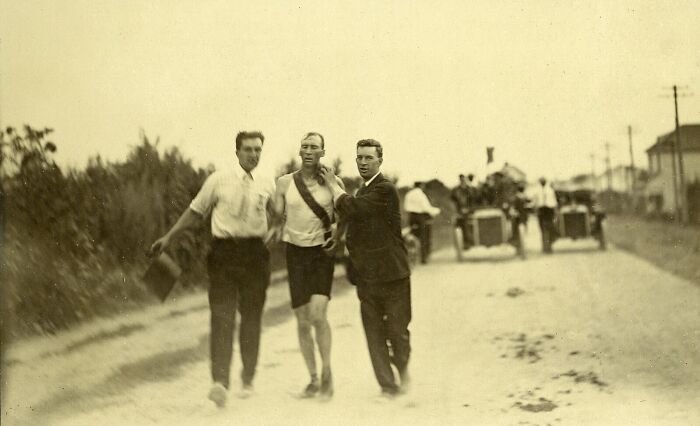Three men during a vintage event from history, walking on a dirt road with early motor vehicles in the background.