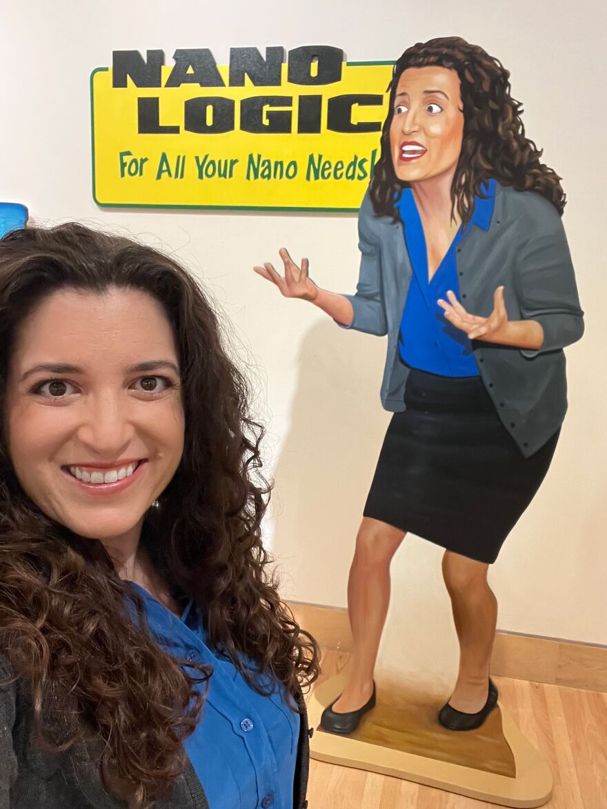 Woman with curly hair smiling in front of a colorful cartoon figure and a bright yellow sign about nano needs