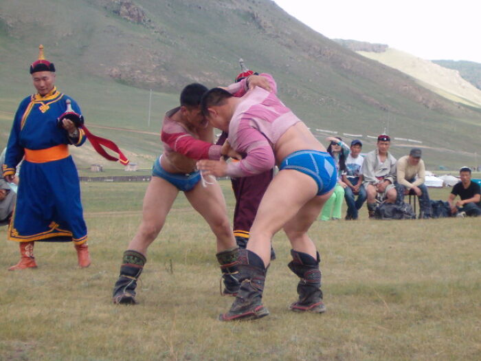 Two men wrestling outdoors in traditional attire with spectators and a referee in a dangerous historical sport setting.