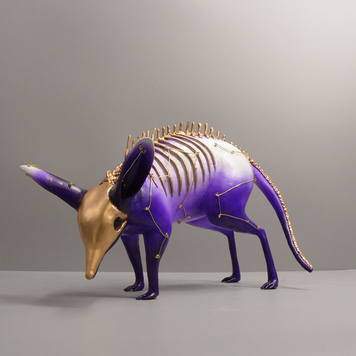 Porcelain sculpture of a mythical creature with gold accents and skeletal details in purple and white tones on a gray surface.