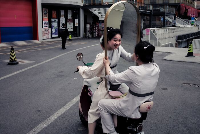 Two women in traditional clothing on a scooter using a mirror, captured in pure street photography awards style.