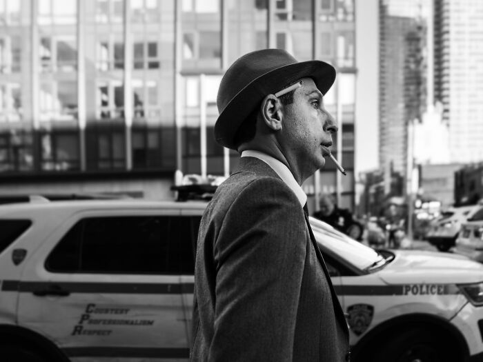 Man in a hat smoking a cigarette on city street with police vehicles in the background, Pure Street Photography Awards style.