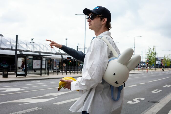 Young man with a bunny-shaped backpack holding bananas, crossing a city street in candid pure street photography style.
