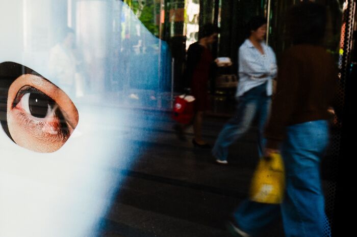 Close-up of an eye through a white mask with blurred pedestrians walking, showcasing street photography capturing urban life.