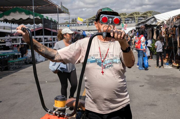 Man wearing red goggles and a graphic tee standing with a custom bike at a street market scene, Pure Street Photography Awards