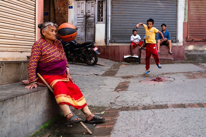 Elderly woman sitting on street curb watching children play basketball in vibrant urban setting, street photography award entry.