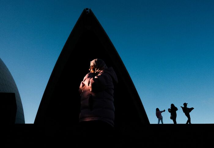 Person in warm clothing lit by sunset light under a triangular structure, showcasing pure street photography awards.