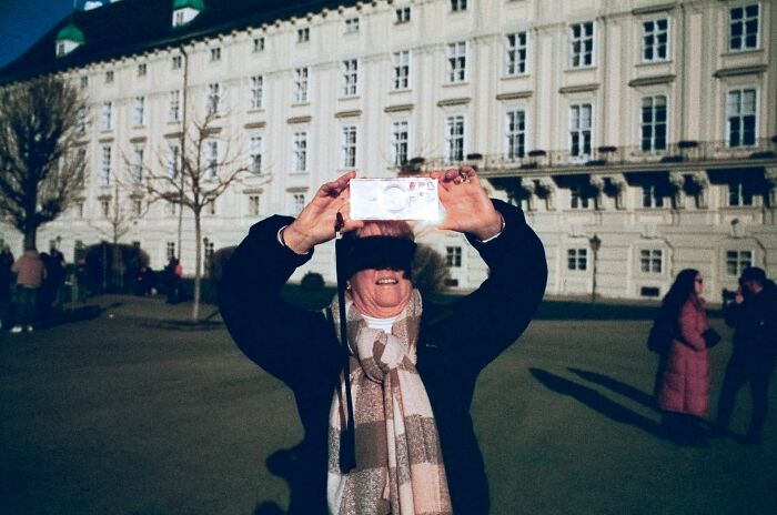 Person in scarf taking a photo outside a historic building, capturing a moment for Pure Street Photography Awards 2025.