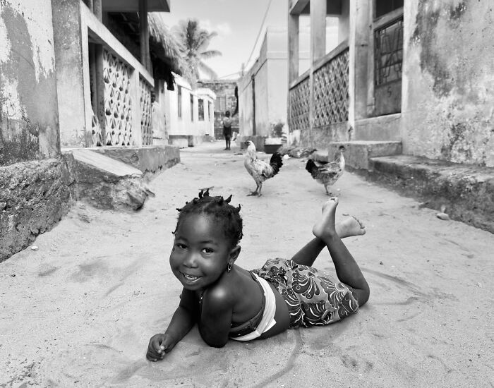Black and white street photography of a smiling child lying on sand with chickens in an urban street scene.
