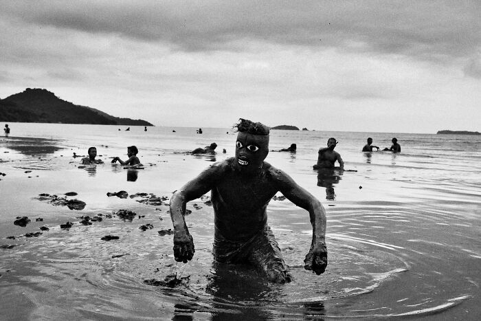 Man covered in mud wearing a mask wading through water while others swim, captured in pure street photography awards.