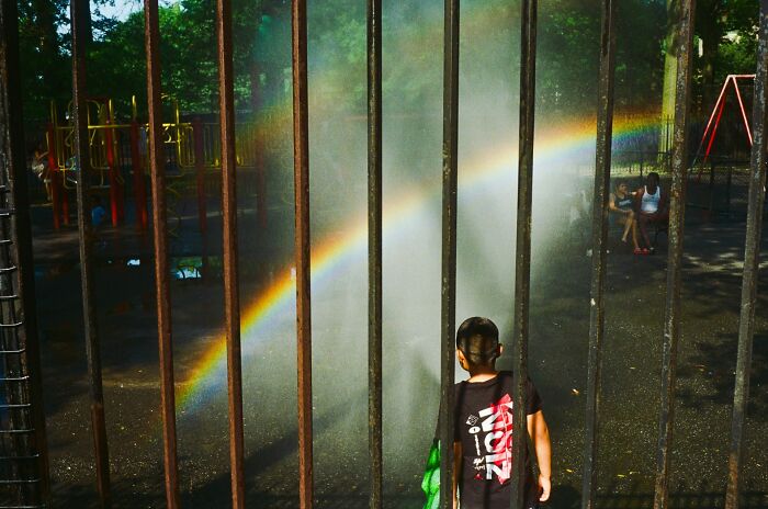 Child playing near a rainbow formed by water spray in an urban playground, capturing the essence of street photography.