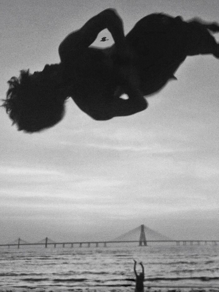 Black and white street photography of a child mid-air above water with a bridge in the background, capturing pure street photography.
