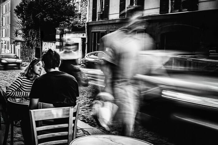 Couple seated at a street café with blurred pedestrians and cars passing by in dynamic street photography.