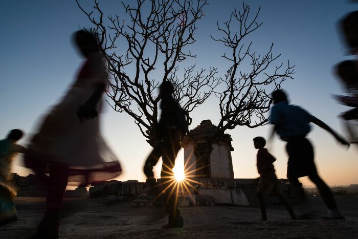 Silhouettes of people walking near a leafless tree and ancient structure at sunset in pure street photography awards.