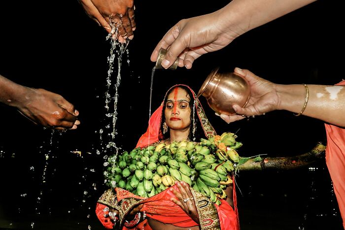Woman in traditional attire with bananas being poured water, captured in pure street photography awards moment.