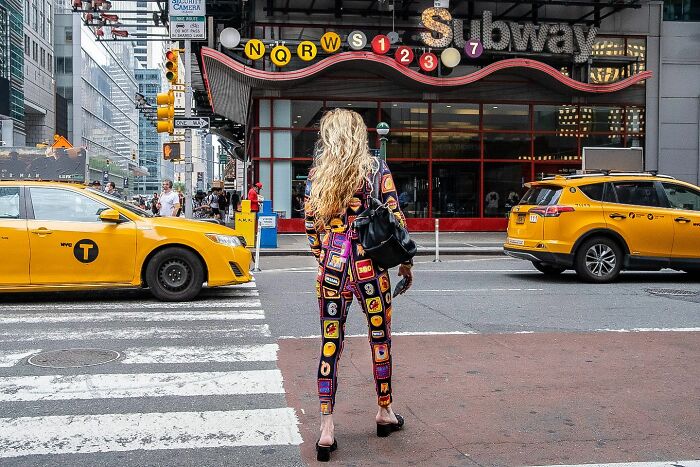 Woman in a colorful outfit standing at a crosswalk near a subway entrance with yellow taxis in busy street photography scene